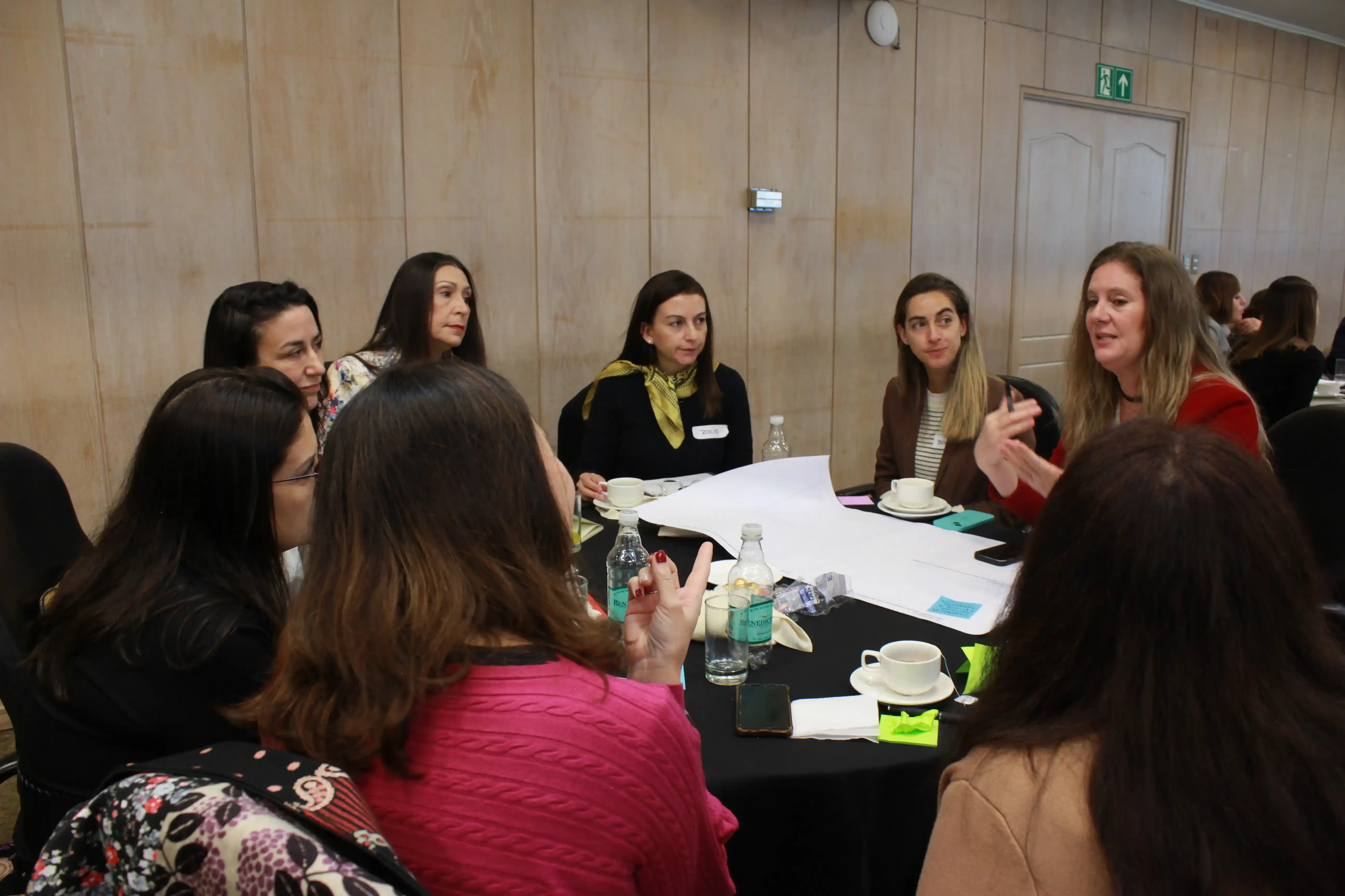 Grupo de participantes conversa alrededor de una mesa mientras una facilitadora guía una actividad con un pliego de trabajo al centro.