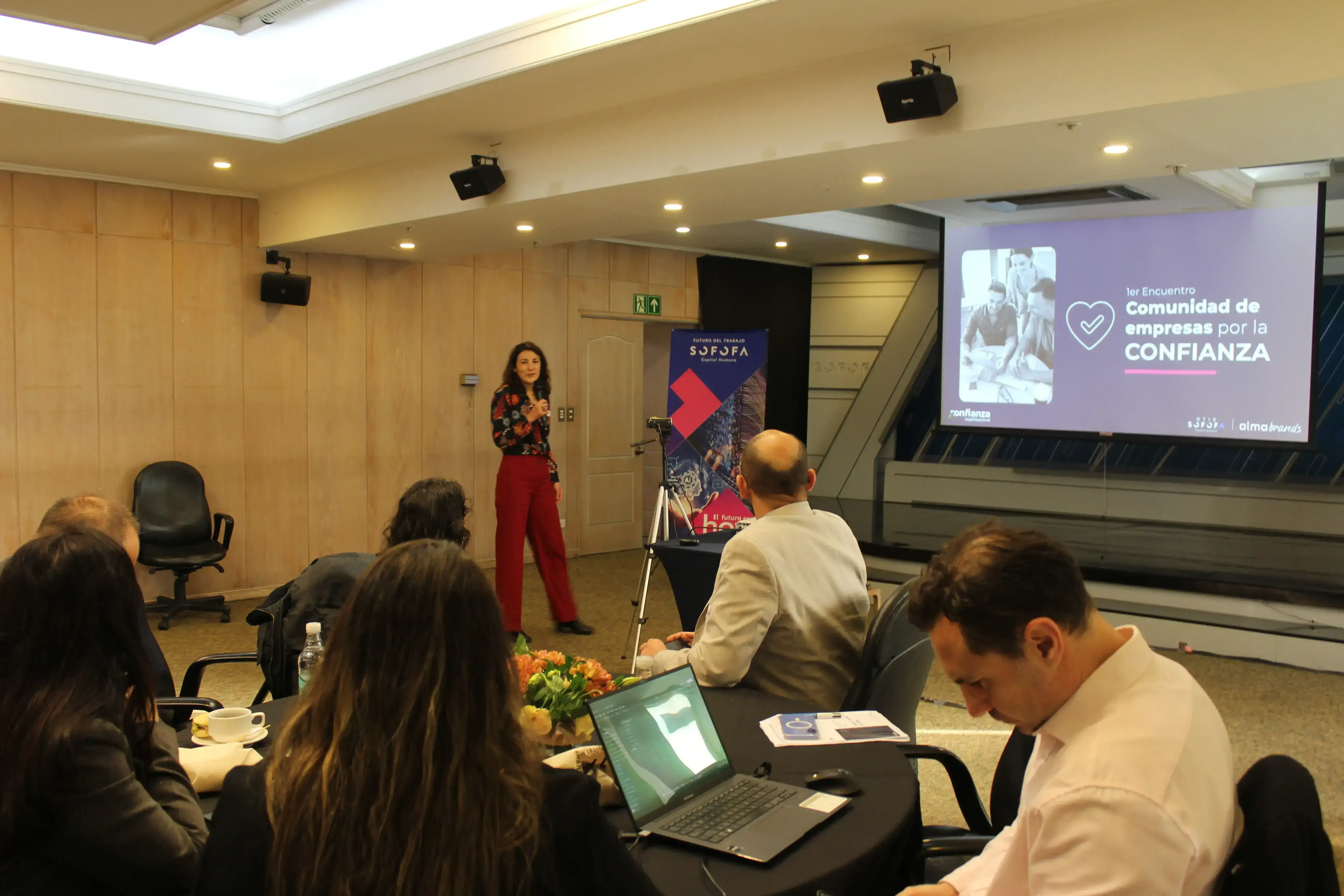 María Teresa Meneses presenta el Primer Encuentro de la Comunidad de Empresas por la Confianza frente a los asistentes y una pantalla con la imagen del evento.