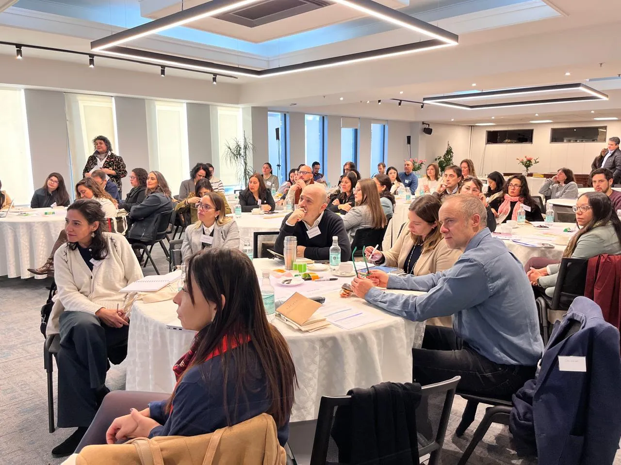 Participantes escuchan atentamente una exposición mientras toman notas desde mesas distribuidas en el salón del encuentro